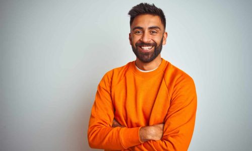Young indian man wearing orange sweater over isolated white background happy face smiling with crossed arms looking at the camera. Positive person.