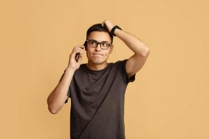 Strange news, problems and difficulties at work. Pensive sad young guy in brown t-shirt and glasses, talking on phone, holding his head and looking up at empty space, isolated on beige background