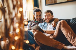 Male couple are lying together on the sofa in their home. One is holding a digital tablet which they are both looking at and using together.